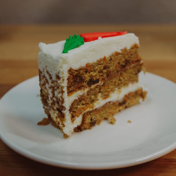A slice of carrot cake with white frosting and decorative green and orange carrots on a white plate.