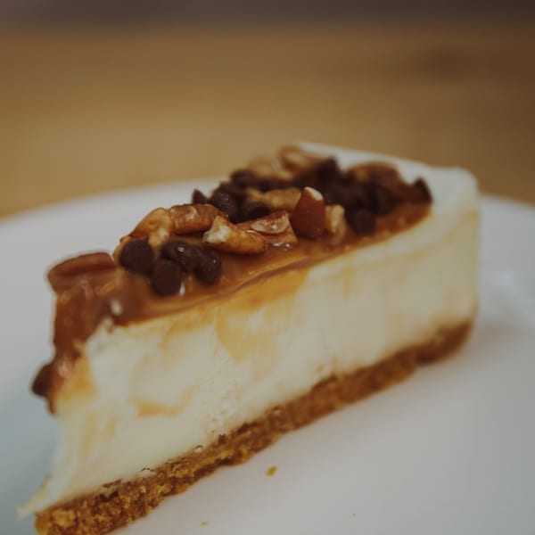 A slice of cheesecake with caramel, chocolate chips, and pecans on a white plate.